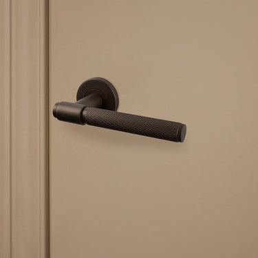 Bronzen Cosenza deurklink met gekartelde structuur op rond rozet op beige deur, rechteraanzicht